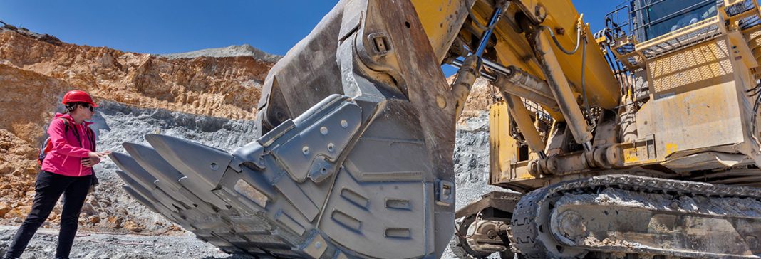 Woman in a red safety hat with red jacket standing in front of an excavator in Open Pit Mine